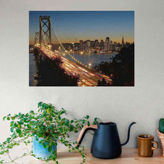Poster of a Golden Gate Bridge and cityscape  at dusk, placed on a wall above a table with a plant and kitchen items.